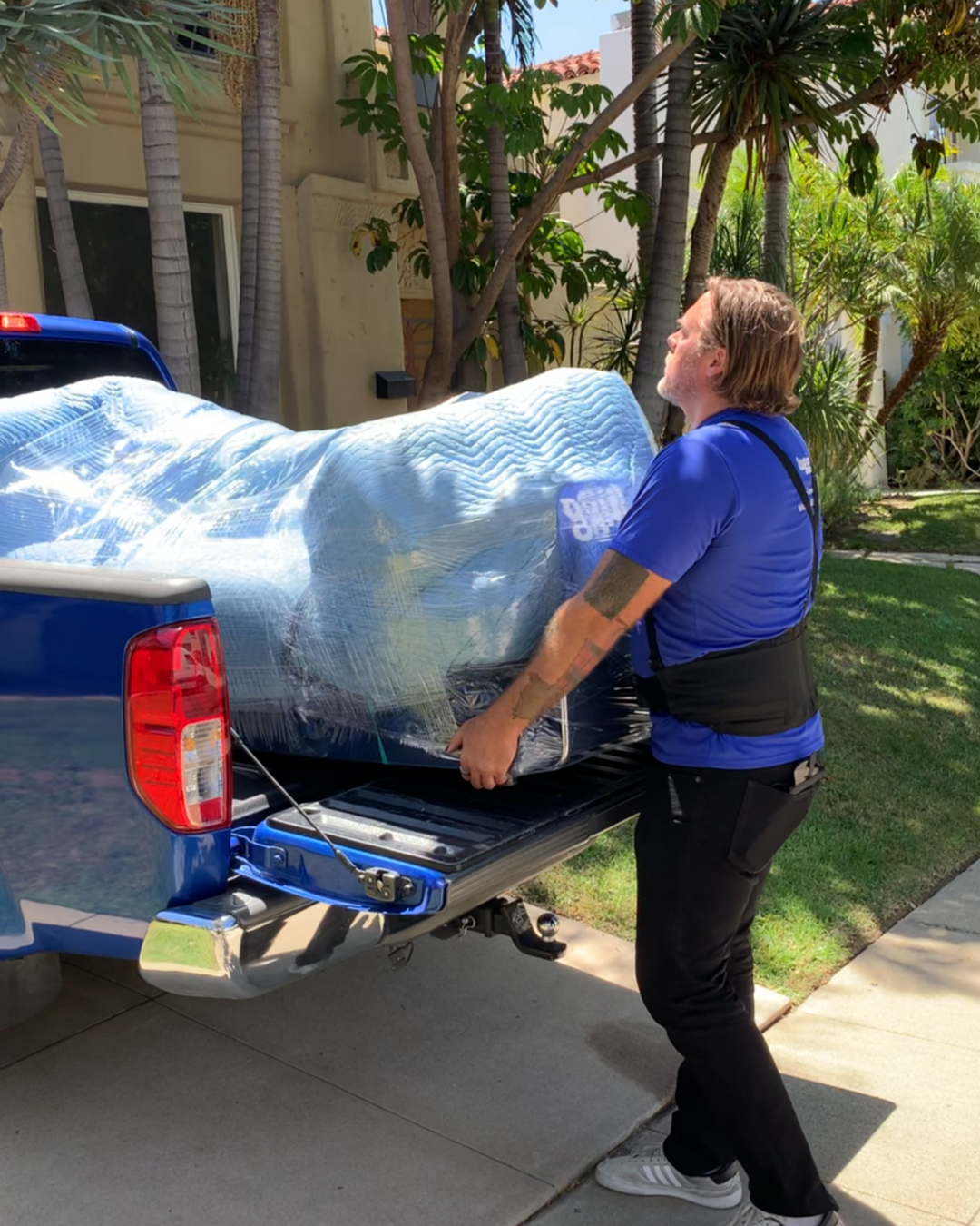 Lugg mover in blue shirt loading a fully wrapped couch into the bed of a blue pickup truck in a residential driveway.