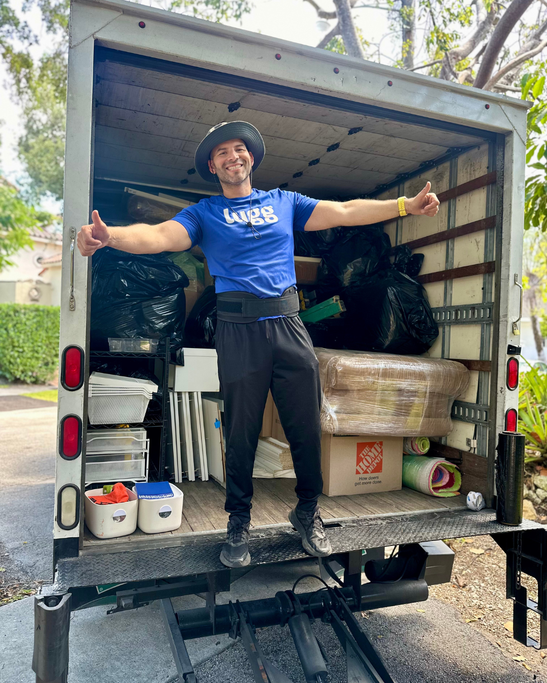 Lugg mover in blue shirt and hat standing with arms outstretched in the back of a fully loaded moving truck.