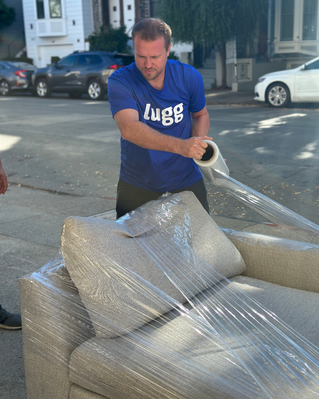 Lugg mover in a blue shirt wrapping a cushioned chair with plastic wrap on a city street.