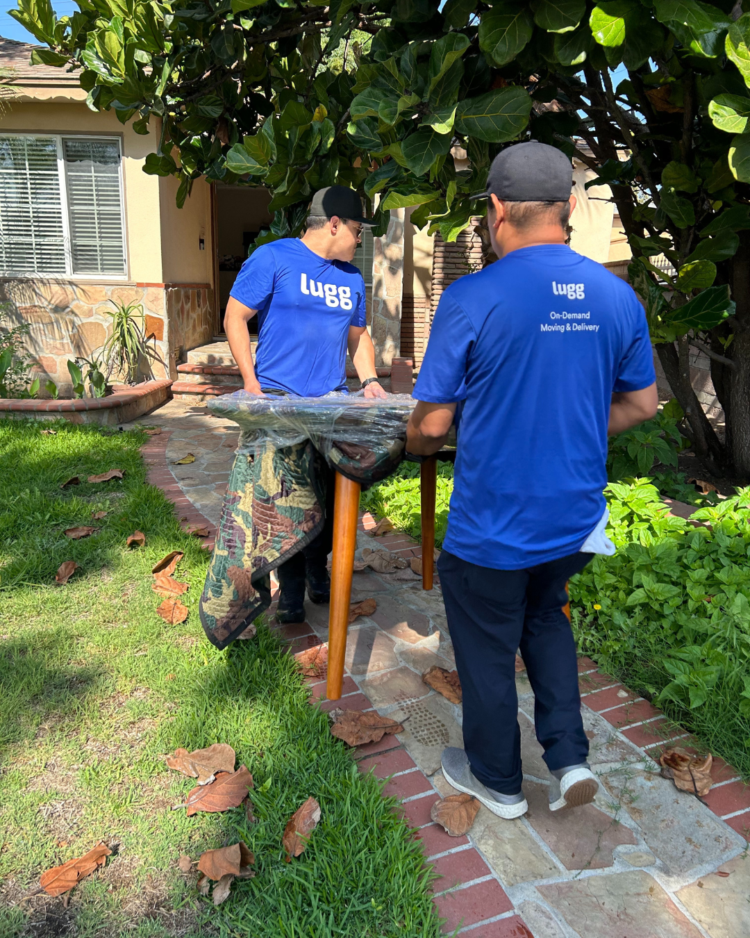 Two Lugg movers in blue shirts carrying a wrapped table along a brick garden path toward a home.