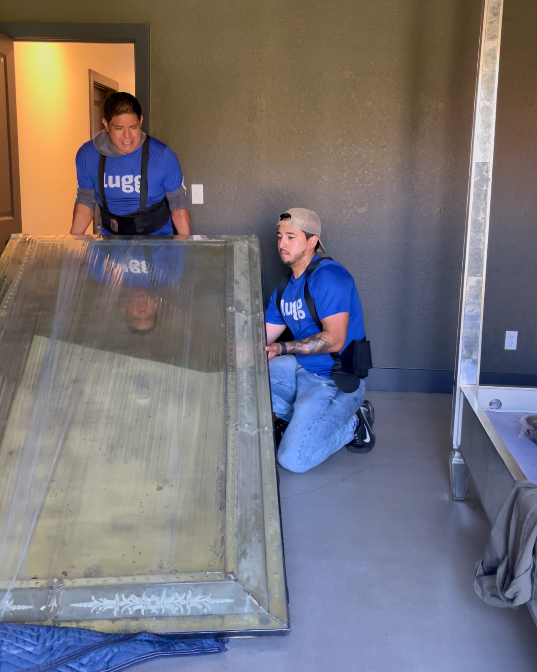 Two Lugg movers lifting and positioning a heavy framed head board indoors, demonstrating safe handling of oversized items.