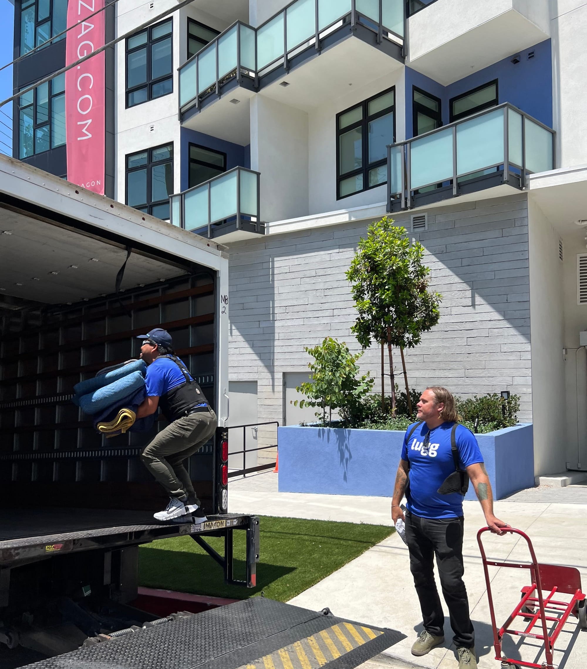 Lugg movers loading supplies into a box truck outside a modern apartment building