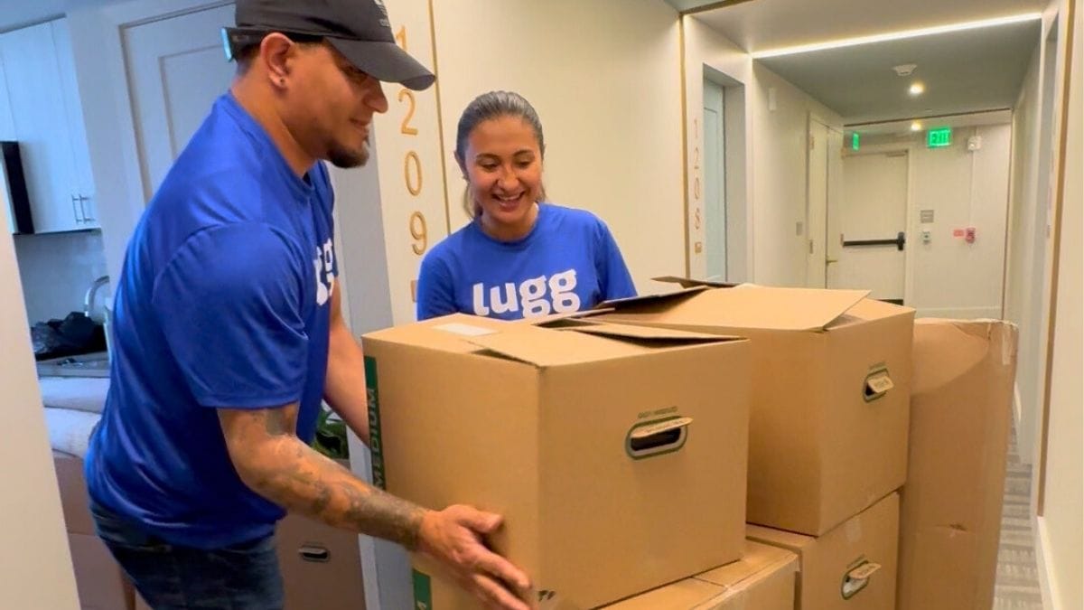 Two Lugg movers in blue shirts carefully carrying stacked moving boxes through an apartment hallway